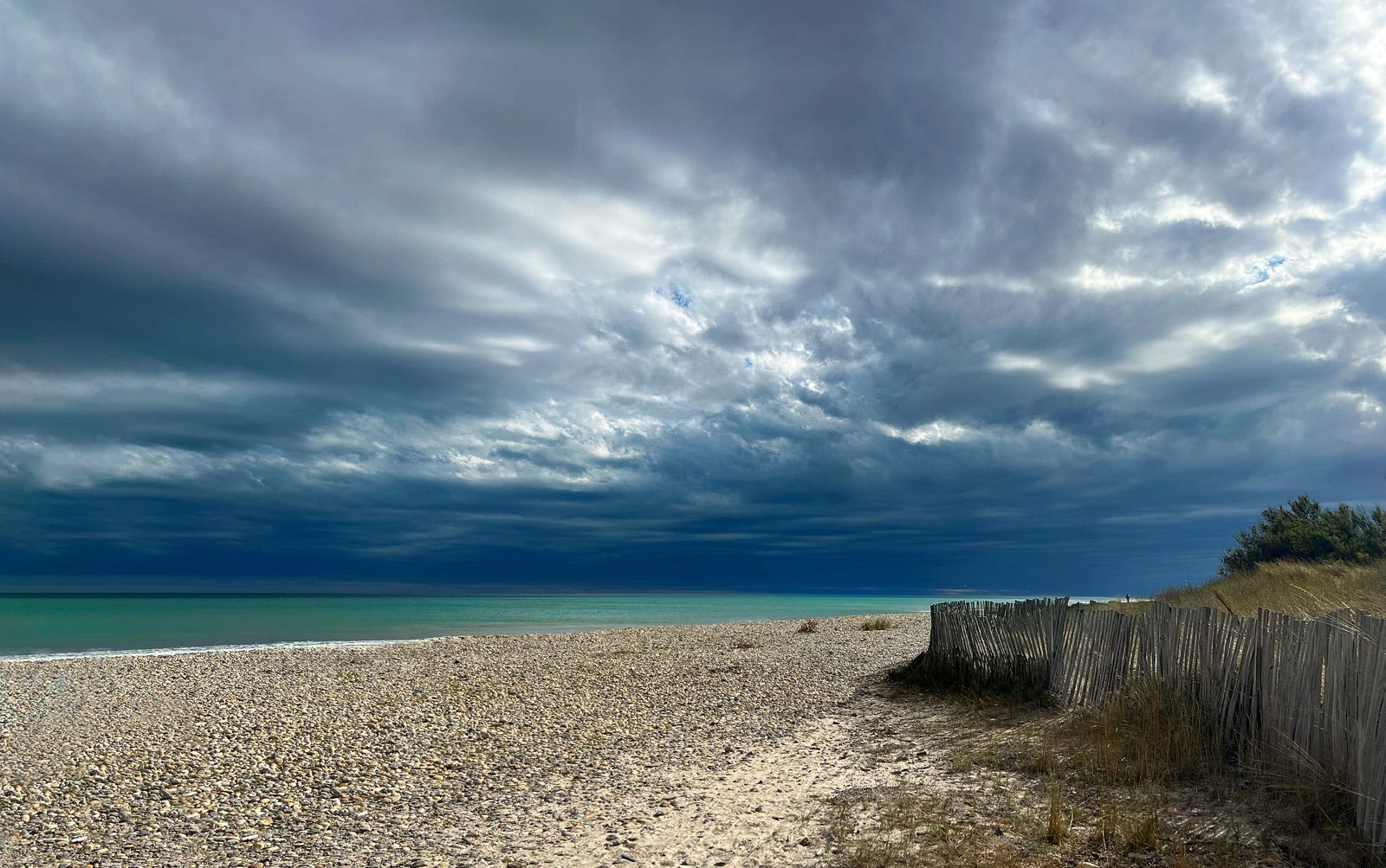 A wide pebble beach stretches toward a calm turquoise sea under a dramatic sky filled with dark, layered storm clouds, with a weathered wooden fence and dune grass along the right side, as light breaks faintly through the clouds, suggesting an approaching or passing storm over a quiet, empty shoreline.
